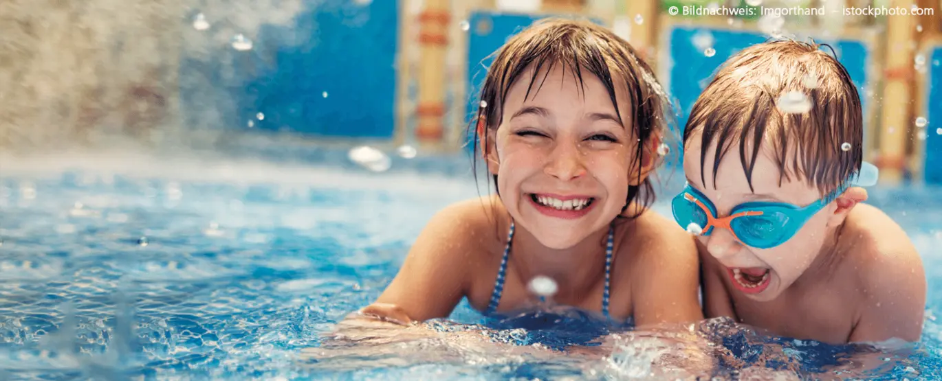 Kampagnenbild zum KVV-Badetag Zwei lachende Kinder, ein Mädchen und ein Junge, etwa sechs Jahre alt, planschen fröhlich im Schwimmbad. Beide tragen eine Taucherbrille und spritzen ausgelassen mit Wasser.