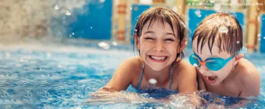 Zwei lachende Kinder, ein Mädchen und ein Junge, etwa sechs Jahre alt, planschen fröhlich im Schwimmbad. Beide tragen eine Taucherbrille und spritzen ausgelassen mit Wasser.