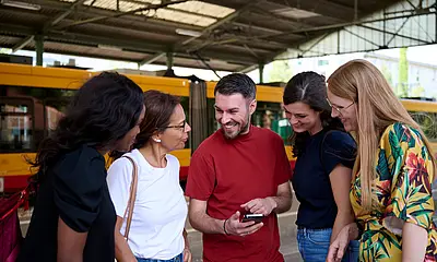Junge Menschen stehen an einer Bahnhaltestelle und schauen gemeinsam auf ein Smartphone.