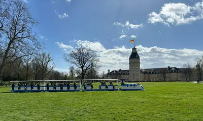 Das Foto zeigt die Schlossgartenbahn mit blau-weißer Porsche-Lok bei der Fahrt durch den Karlsruher Schlossgarten. Im Hintergrund ist ds Karlsruher Schloss zu sehen.