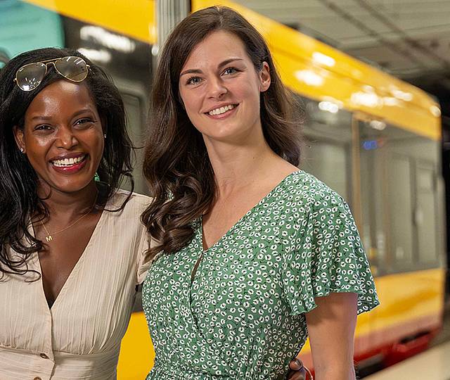 Zwei junge Frauen stehen vor einer Bahn an einer unterirdischen Haltestelle.