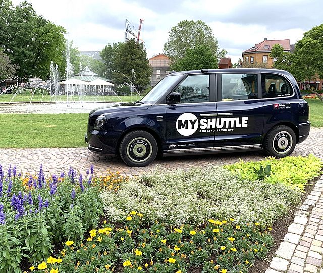 Ein  London Taxi mit der MyShuttle Beklebung steht an einem schönen Platz mit Springbrunnen.