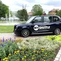 Ein  London Taxi mit der MyShuttle Beklebung steht an einem schönen Platz mit Springbrunnen.