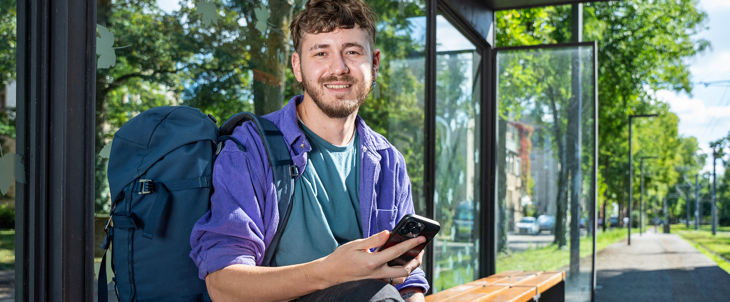 Ein junger Mann sitzt lächelnd an der Haltestelle und hält sein Smartphone.