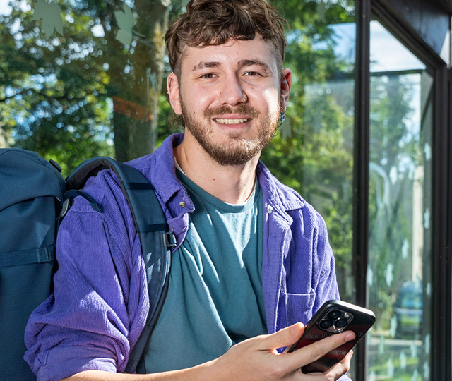 Ein junger Mann sitzt lächelnd an der Haltestelle und hält sein Smartphone.