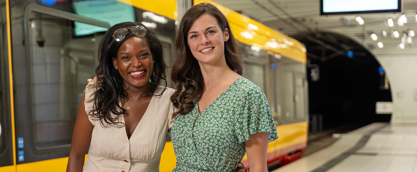 Zwei junge Frauen stehen vor einer Bahn an einer unterirdischen Haltestelle.