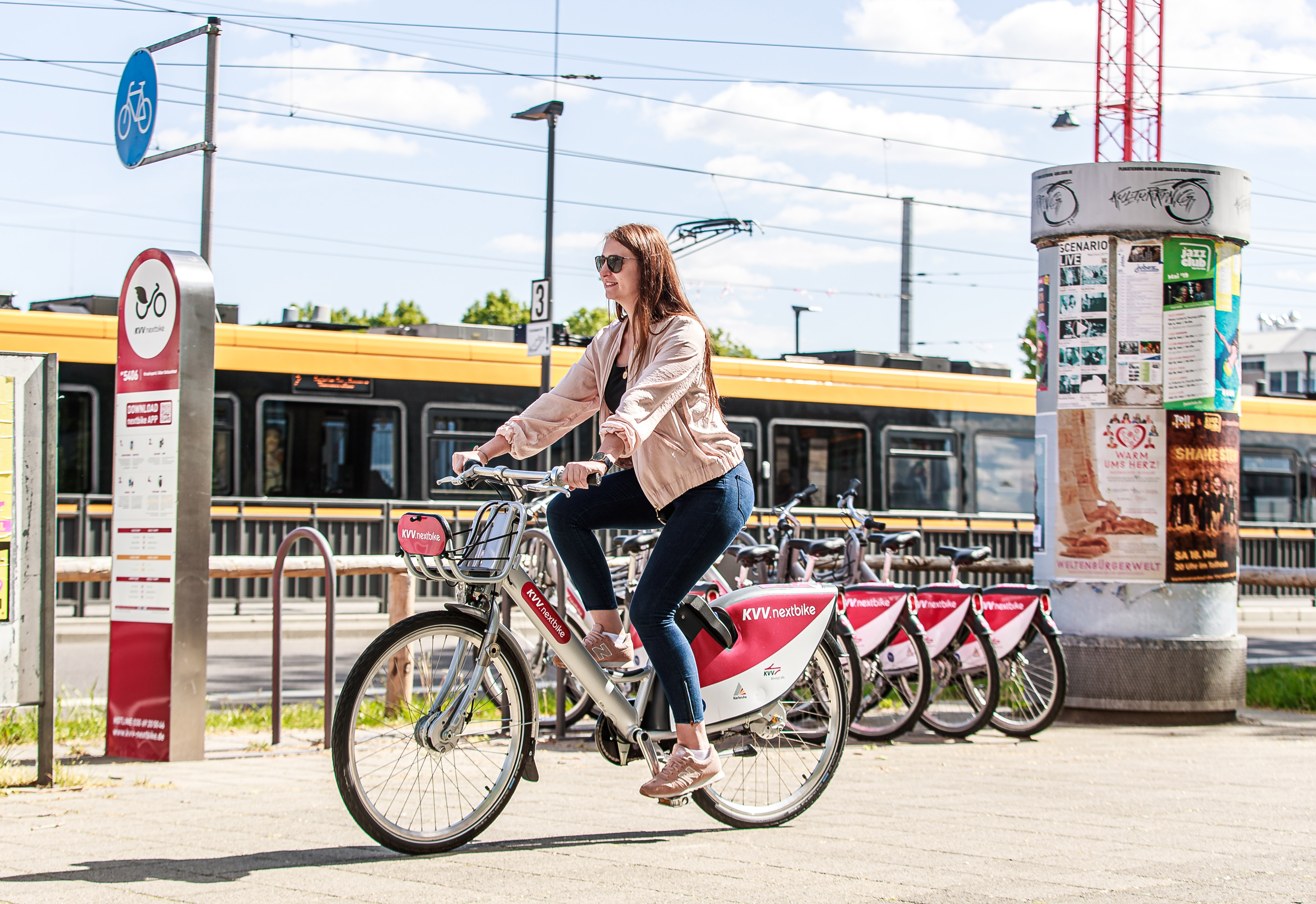 Brunette Frau fährt auf einem Fahrrad der Firma nextbike. Im Hintergrund sind weitere Fahrräder und eine gelbe Straßenbahn zu sehen. Der Himmel ist blau und leicht bewölkt.